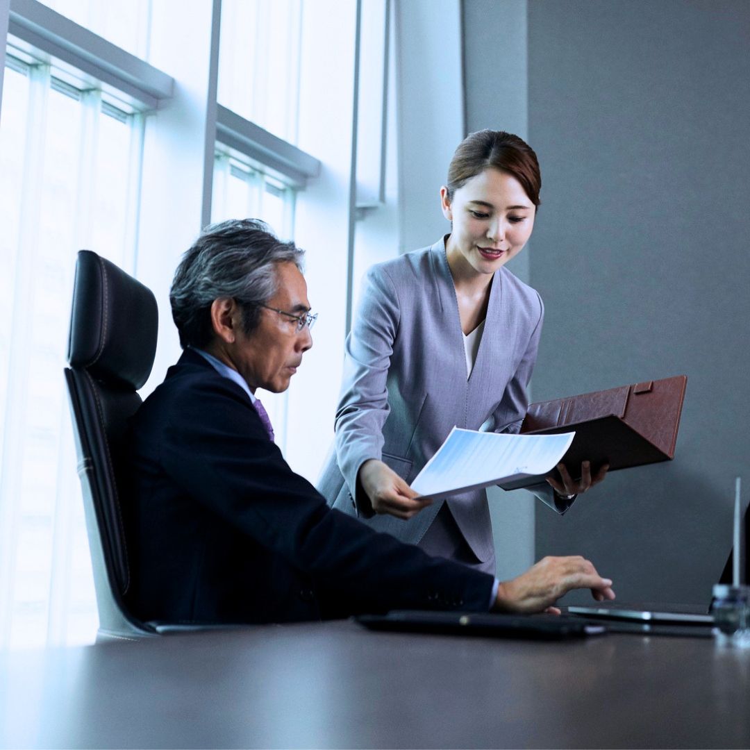 Executive assistant in business attire briefing a senior executive seated at his desk, illustrating the trusted role of EAs and chiefs of staff.
